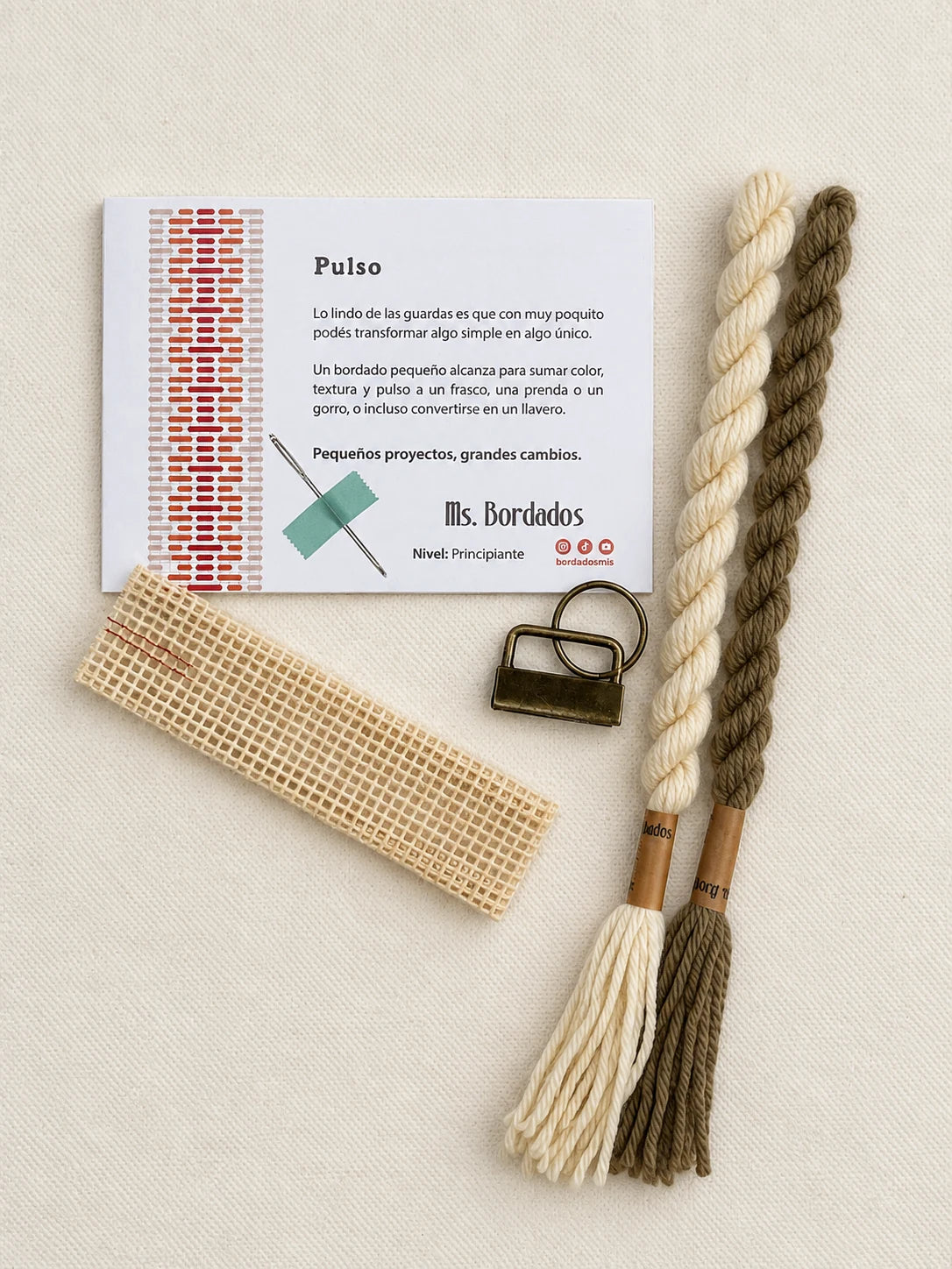 Componentes del Kit Franja Pulso Lino (formato Llavero): cañamazo, lanas 100% uruguayas en Crudo y Topo, aguja, manual impreso y herraje del llavero — Ms. Bordados
